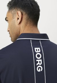 Vyras su trumpais garbanotais plaukais, vilkintis tamsiai mėlyną polo marškinėlį su baltais kraštais ir užrašu "BORG" vertikaliai ant nugaros apykaklės juostos.