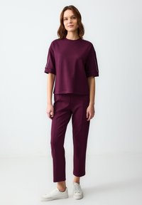 Bordeaux gekleed set bestaande uit een boxy top met korte mouwen en slim-fit broeken. Zacht materiaal met opgerolde mouwen en subtiele zijaccenten. Witte sneakers.
