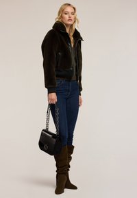 Nainen, jolla on päällään tummanruskea turkitakki, siniset farkut ja rennot ruskeat saappaat, pitää mustaa käsilaukkua, jossa on ketjuhihna, ja seisoo tasaisella taustalla.