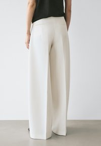Personne portant un pantalon blanc taille haute à jambes larges et un haut noir sans manches, debout devant un fond clair uni.