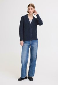 Cardigan en maille côtelée marine avec un col en V, fermeture par boutons, et manches longues, associé à un jean bleu clair à jambes larges et des chaussures noires.