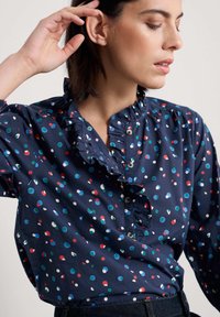 Navy blauwe blouse met een gerimpelde kraag, versierd met multicolored stippen. Knopen zichtbaar bij de hals; katoenen stof, casual pasvorm.