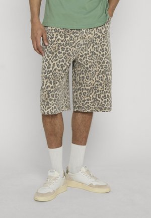 Mand iført leopardmønstrede shorts i midterlængde, hvide crew-sokker, hvide sneakers og en grøn skjorte med hånden i lommen.