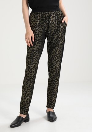 Person trägt schwarze und goldene Leopardenmuster-Hose mit elastischem Bund und schwarze Slipper, steht mit einer Hand in der Tasche.