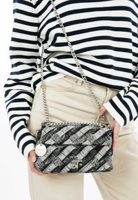 Borsa a tracolla nera e grigia trapuntata con catena argentata, caratterizzata da un logo e un tag rotondo. Texture liscia con un motivo geometrico.