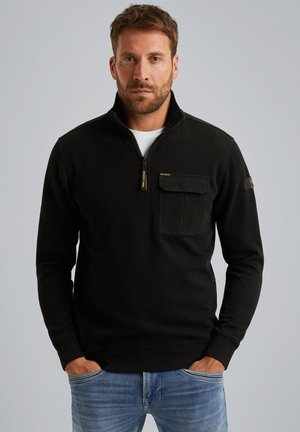 Sort zip-neck pullover med ribbet tekstur, der har en frontlomme og logoaccent. Båret med blå jeans, er pasformen afslappet og uformel.