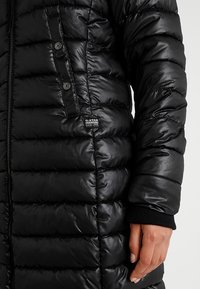 Manteau doudoune noir avec coutures horizontales, fermeture éclair à l'avant et poche boutonnée. Présente des poignets élastiques et un petit logo de marque sur le côté.