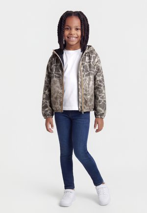 Naeratav tüdruk, kellel on patsiga juuksed ning kes kannab mustrilist halli kapuutsiga jopet, valget särki, siniseid pükse ja valgeid tenniseid, seisab valgel taustal.