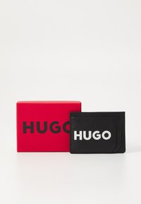 Portafoglio in pelle nera con logo "HUGO" in bianco impresso, accanto a una scatola rossa con lo stesso logo in testo nero in grassetto.