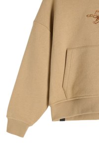 Beżowy sweter z miękkiej tkanki, w oversize'owym kroju, z prążkowanymi mankietami i dołem. Posiada kieszeń z przodu oraz subtelny wytłoczony detal z logo.