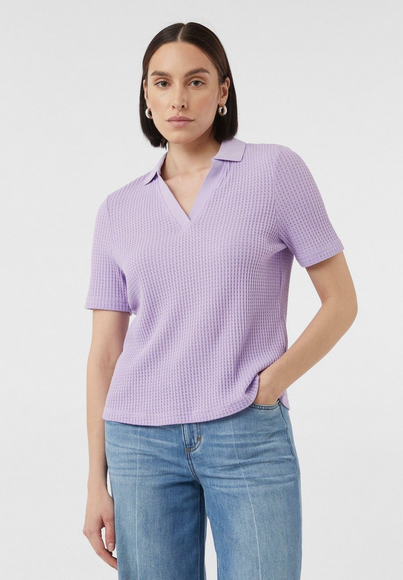 Helllila kurzärmliges Poloshirt mit Waffelstruktur und V-Ausschnitt. Kombiniert mit blauen Jeans, die Hände in den Taschen.