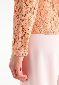 Perzikkleurige kantblouse met bloemmotief, lange mouwen en een gestructureerde afwerking, gecombineerd met een gladde roze stof erachter.