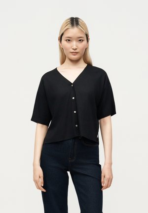 Kobieta o prostych blond włosach, ubrana w czarną koszulę z krótkim rękawem zapinaną na guziki i ciemne dżinsy, stojąca na jasnym jednolitym tle.