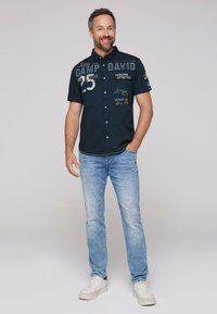Navy short-sleeve button-up shirt with white graphics, paired with light blue denim jeans and white sneakers.