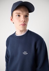 Ναυτικό φούτερ με λευκό λογότυπο στο στήθος, στρογγυλή λαιμόκοψη, πλέξη στους καρπούς. Συνδυασμένο με σκούρο μπλε καπέλο, με υφασμάτινη γείσο.