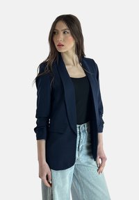 Blazer navy dal taglio slim, con apertura frontale e spalle strutturate. Tessuto morbido e maniche a tre quarti. Abbinato a jeans a gamba larga in denim chiaro.