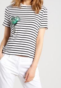 Černobílé pruhované tričko s krátkým rukávem, na levé straně s zelenou vyšitou ptáčkem, nošené s bílými kalhotami.