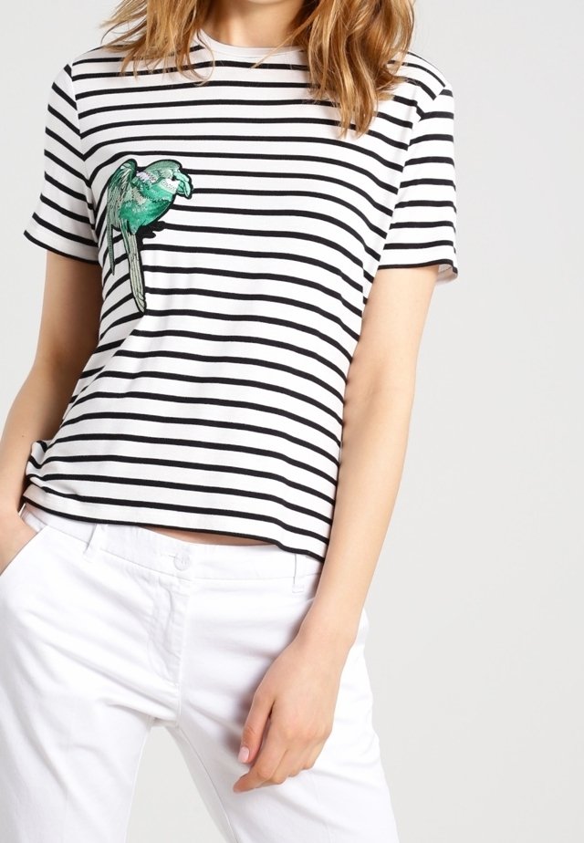 Černobílé pruhované tričko s krátkým rukávem, na levé straně s zelenou vyšitou ptáčkem, nošené s bílými kalhotami.