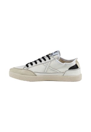 Zapatilla blanca de corte bajo con cordones negros, suela beige y detalle negro en el talón, mostrada de lado sobre un fondo blanco.