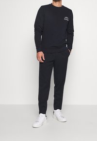 Sudadera de marinero con cuello redondo y logo en el pecho, combinada con pantalones oscuros de rayas y zapatillas blancas para un look casual.