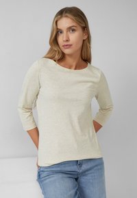 Light beige 3/4 sleeve top with a rounded neckline, made from soft fabric, featuring a smooth texture and a relaxed fit. Worn with blue jeans.