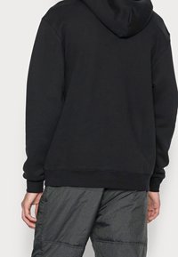 Felpa nera con cappuccio, tasca a marsupio e orlo a coste. Il tessuto appare morbido e strutturato. Abbinata a pantaloni chiari e textured.