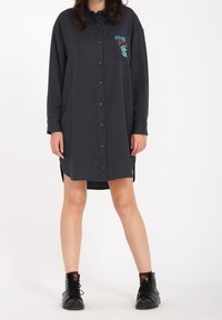 Robe chemise noire à manches longues, avec une poche poitrine ornée d'un graphique coloré et un ourlet légèrement incurvé. Associée à des bottines noires.