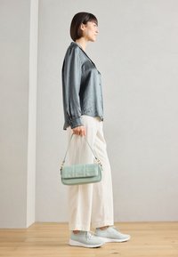 Woman in light blue satin blouse, cream wide pants, and white sneakers holding a mint green handbag in a neutral indoor setting.