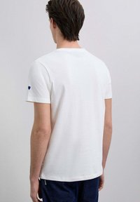 Camiseta blanca de manga corta con escote redondo, con un pequeño detalle de corazón azul en la manga izquierda. Confeccionada con una tela suave.