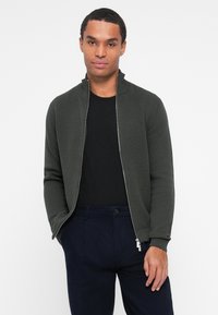 Sweat-shirt zippé vert foncé avec un motif en tricot texturé, col montant et poignets ajustés, associé à un t-shirt noir et un pantalon bleu marine.