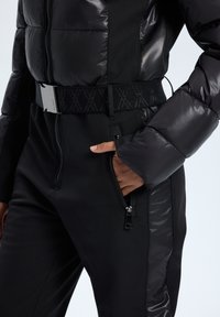 Czarna kurtka puchowa z pikowanym wzorem i błyszczącym wykończeniem, zestawiona z dopasowanymi czarnymi spodniami z zamkami błyskawicznymi i wzorzystym elastycznym paskiem.