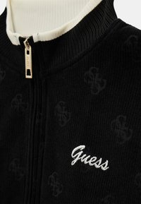 Veste zippée noire avec col côtelé et motif logo subtil. Présente une broderie blanche "Guess" et des détails en métal zippés.