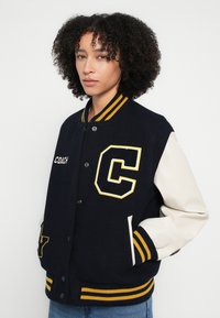 Tengerészkék és krémszínű egyetemi dzseki sárga díszítésekkel, amelyen kiemelkedő "C" logó, bordázott gallér és csíkos mandzsetták találhatók. Elől gombos záródás.