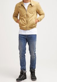 Tan bomberjakke med lynlås foran, elastiske manchetter og kant, båret over en hvid skjorte, parret med blå jeans og sorte høj-top støvler.