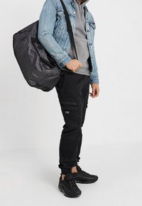 Henkilö, jolla on päällään mustat cargo-housut, mustat lenkkarit, harmaa huppari ja sininen farkkutakki, kantaa iso musta matkakassi toisen olkapään yli.