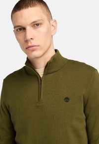 Olivgrüner gestrickter Pullover mit hohem Kragen und halbem Reißverschluss. Besitzt dezente horizontale Rippungen und ein kleines schwarzes Logo auf der Brust.