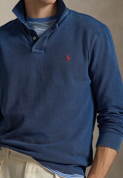 Navy blue long-sleeve polo shirt with a ribbed collar and red logo accent. Underneath, a striped shirt in blue and white is visible.