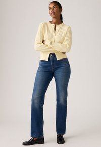 Καρντιγκάν σε ανοιχτό κίτρινο με πλεκτή υφή, στρογγυλή λαιμόκοψη και κούμπωμα με κουμπιά, συνδυασμένο με μπλε τζιν παντελόνι ίσιας γραμμής και μαύρα παπούτσια.