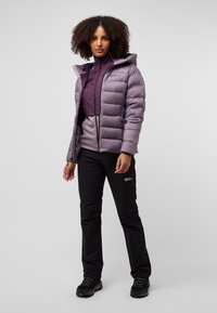 Giacca a vento viola con cuciture orizzontali, chiusura con zip e cappuccio; indossata sopra un top viola; pantaloni e calzature neri; vestibilità snella.