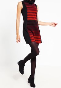 Knielanges, ärmelloses Kleid in Schwarz und roten Streifen, mit hohem Kragen und taillierter Passform, kombiniert mit schwarzen Strumpfhosen und Stiefeletten.