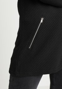 Svart texturerad jacka med en diagonal ficka på sidan, tillverkad av ett mysigt material med ett subtilt mönster.