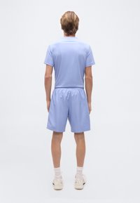 Άτομο που στέκεται προβάλλοντας την πλάτη, φορά ει φωτεινό μπλε μπλουζάκι, σορτς που ταιριάζει, λευκές κάλτσες και λευκά αθλητικά παπούτσια σε απλό φόντο.