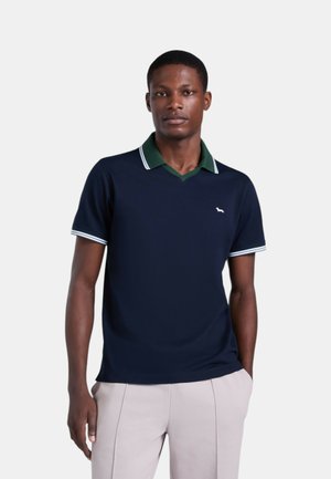 Uomo che indossa una polo blu navy a maniche corte con colletto verde e rifiniture bianche, abbinata a pantaloni beige chiaro, in piedi davanti a uno sfondo semplice.