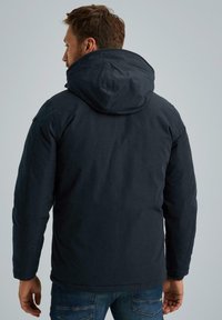 Giacca con cappuccio blu navy dal tessuto liscio, dotata di chiusura con zip e tasche laterali. Il retro presenta un taglio leggermente allungato.