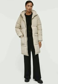 Beige pufferjacka med huva, quiltat mönster, framdragkedja och sidofickor. Bärs över en mörk stickad topp och svarta vida byxor.