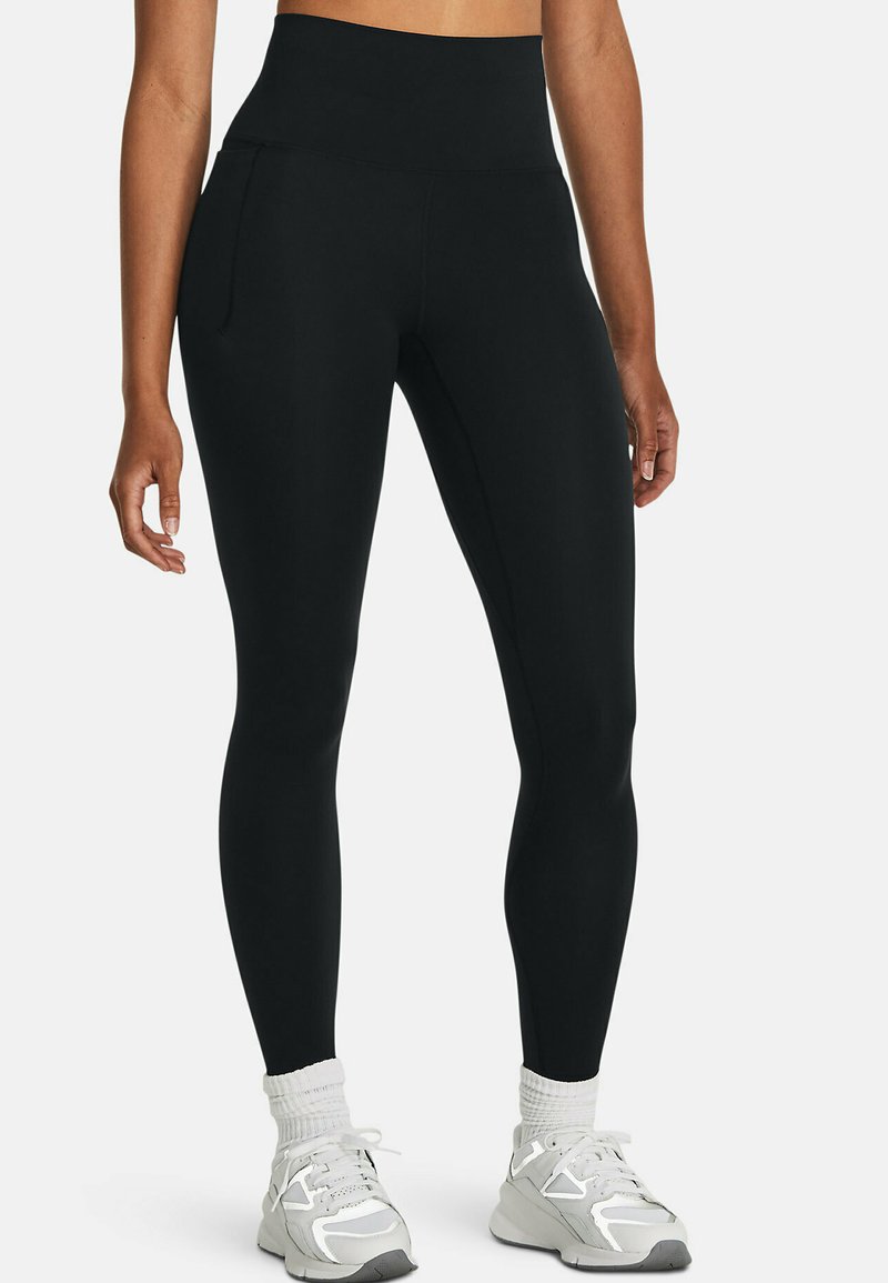 Vrouw in zwarte high-waisted leggings met zijzakken, witte sokken en witte sportschoenen, staande tegen een effen achtergrond.