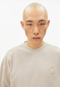 Beige Sweatshirt mit erhabenem Logo auf der Brust, rundem Ausschnitt und sichtbaren Nahtdetails. Glatte Textur und lockere Passform.