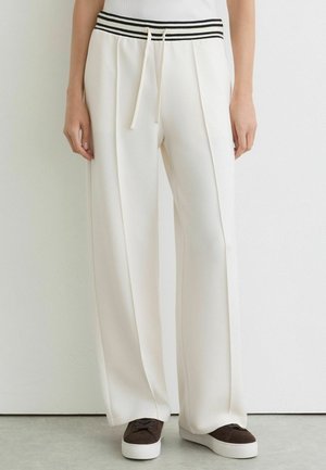 Persoon draagt crème wijde broek met hoge taille, zwart-witte gestreepte tailleband en beige trekkoord, gecombineerd met bruine sneakers met witte zolen.