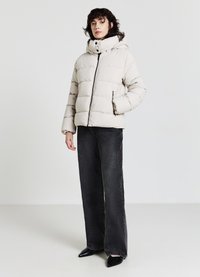 Piumino color beige chiaro con zip frontale, cappuccio ampio, polsini elastici e due tasche laterali. Indossato con jeans neri a vita alta.