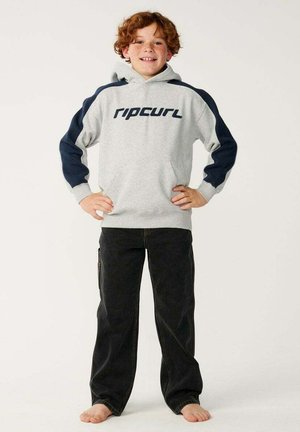 Niño sonriente pelirrojo está descalzo con las manos en las caderas, vistiendo una sudadera con capucha gris Rip Curl con mangas azul marino y pantalones negros sobre un fondo liso y claro.